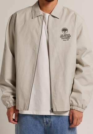 Homme portant une veste beige zippée avec l'inscription "Los Angeles Griffith Park California" et un logo d'arbre sur le côté gauche de la poitrine, un t-shirt blanc, et un jean bleu.