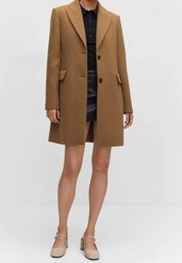 Bruine wolmix jas met een getailleerd ontwerp, geknipte revers, twee voorzakken en twee knopen, gedragen over een zwarte polo en leren rok.