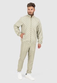 Helle khakifarbene Jacke mit geometrischem Muster, Reißverschluss vorne und elastischen Bündchen, kombiniert mit passenden Hosen und weißen Sneakern.