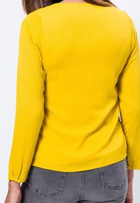 Gul langærmet top med rund halsudskæring, blød tekstur og rynkede manchetter, kombineret med grå jeans med baglommer.