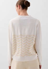 Creamfarvet striktrøje med et tekstureret zigzag mønster i lys gul på ryggen. Ribbetræk ved håndleddene og kanten, afslappet pasform og subtile syninger.