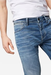 Los jeans de mezclilla azul claro presentan un ajuste slim, cinco bolsillos y un ligero desgaste. La tela tiene una textura suave con un ligero estiramiento.