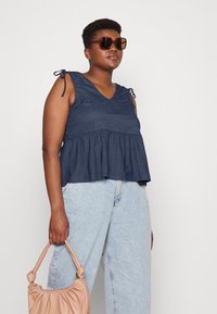 Top peplum azul marinho com decote em V e alças com laço, combinado com jeans de ganga azul clara e uma mala rosa suave.