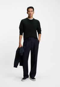 Czarny sweter o teksturowanej powierzchni z okrągłym dekoltem, zestawiony z granatowymi spodniami o szerokich nogawkach. Trzyma granatową kurtkę i ma na sobie czarne sneakersy.