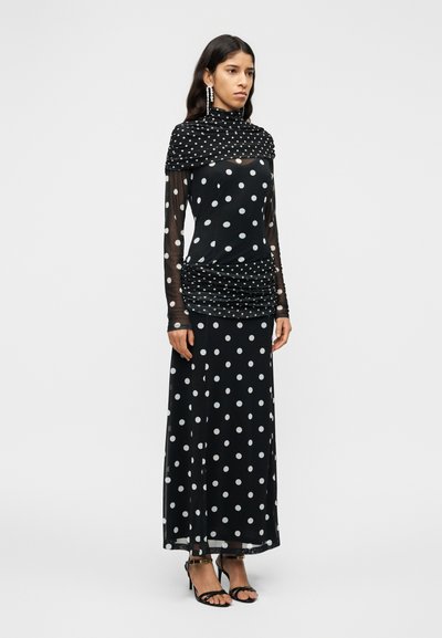 Μαύρο μακρύ φόρεμα με πουά σχέδιο, διάφανα μακριά μανίκια και σούρες στο μπούστο. Διαθέτει ψηλό λαιμόκοψη και φαρδύ, ρέοντα μπροστινό μέρος. Μαύρα πέδιλα με λουράκια.