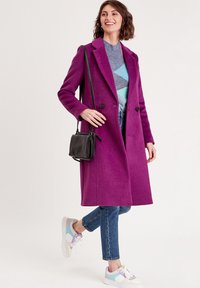 Manteau en mélange de laine violet avec un design croisé, boutons noirs, associé à un pull bleu à motifs, un jean et des baskets multicolores.