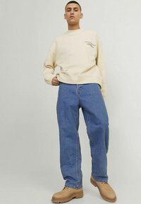 Sweatshirt bege clara com decote redondo e pequeno detalhe em texto, combinada com jeans de ganga azul soltos e botas de trabalho castanhas.