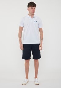 Polo bianco con logo navy sul petto, abbinato a pantaloni corti blu scuro. Il modello indossa sneakers bianche, in una posa neutra.