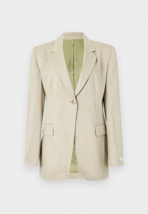 Femme debout, une main dans la poche d’un pantalon rose taille haute, portant un blazer beige, un top blanc, des boucles d’oreilles créoles et des ballerines pointues beiges.