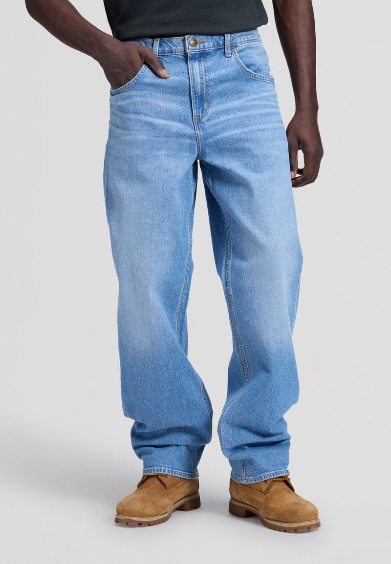 Homme portant un jean bleu clair ample et des bottes de travail couleur beige, une main dans la poche, sur un fond uni.