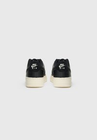 Une paire de baskets noires Nike Air avec des semelles blanches, vue de l'arrière sur un fond gris clair uni.