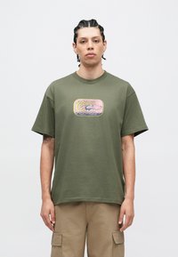 Olívazöld pamut póló laza szabással, színes grafikával az elején, rövid ujjú és kerek nyakú.