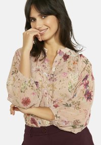 Ziedu blūze caurspīdīgā rozā audumā ar pogām priekšpusē, pūkainām piedurknēm un dažādiem krāsainiem ziedu rakstiem visā audumā.