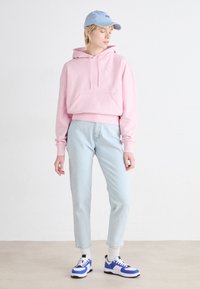 Sudadera rosa clara con un bolsillo frontal, combinada con jeans azul claro. Lleva una gorra azul clara y zapatillas blancas con acentos azules.