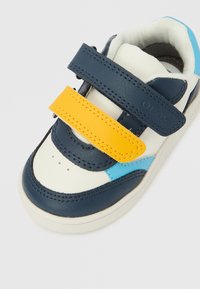 Pantof sport cu exterior din piele bleumarin, accente albe, detalii albastre și o curea velcro galbenă. Prezintă perforații pe partea superioară a degetelor.