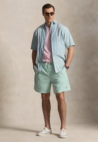 Άνδρας με γυαλιά ηλίου, ανοιχτό μπλε κοντομάνικο πουκάμισο πάνω από ροζ μπλούζα, πράσινες βερμούδες και άσπρα αθλητικά παπούτσια, στέκεται με τα χέρια στις τσέπες.