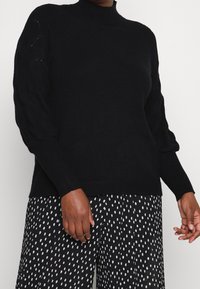 Schwarzer Pullover mit hohem Kragen und gestricktem Zopfstrickmuster an den Ärmeln. Weiche Textur mit gerippten Bündchen und Saum. Kombiniert mit gemusterten schwarzen Hosen.