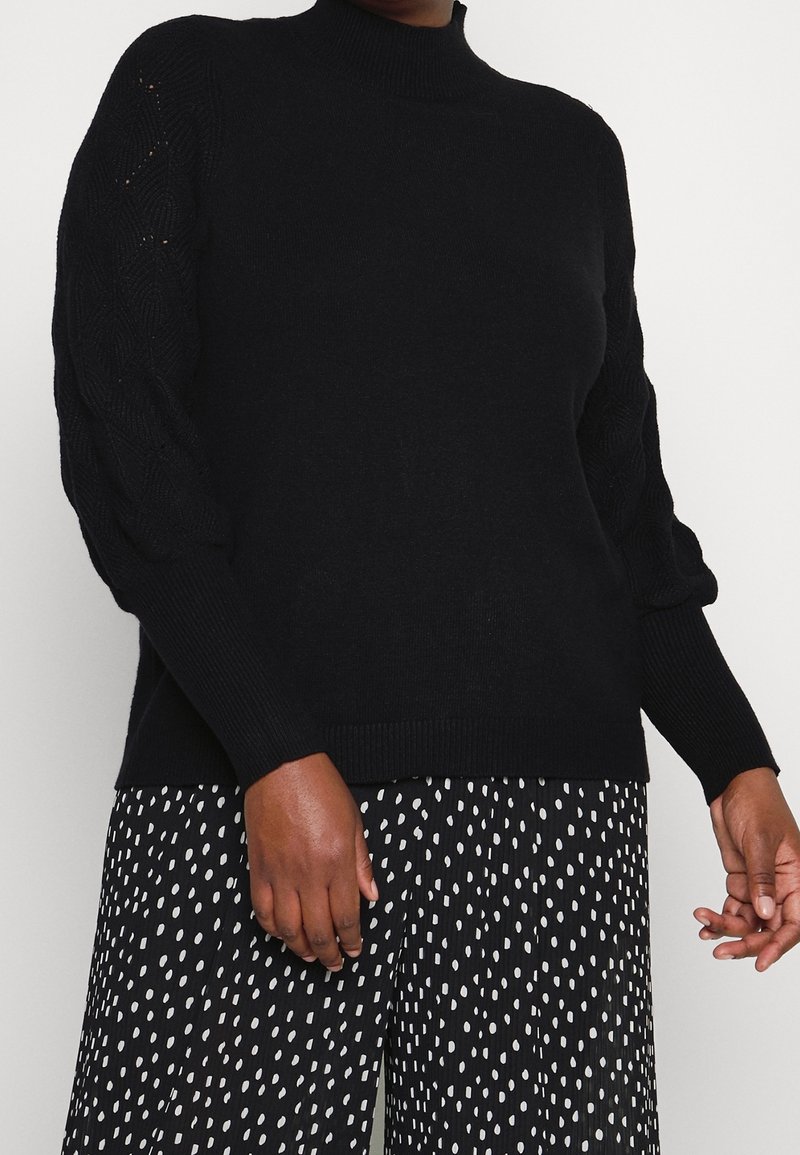 Schwarzer Pullover mit hohem Kragen und gestricktem Zopfstrickmuster an den Ärmeln. Weiche Textur mit gerippten Bündchen und Saum. Kombiniert mit gemusterten schwarzen Hosen.