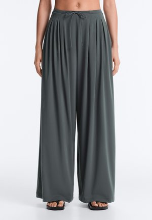 Pantalon gris foncé à jambes larges avec plis et taille à cordon, porté avec des sandales noires, montré de la taille aux pieds.