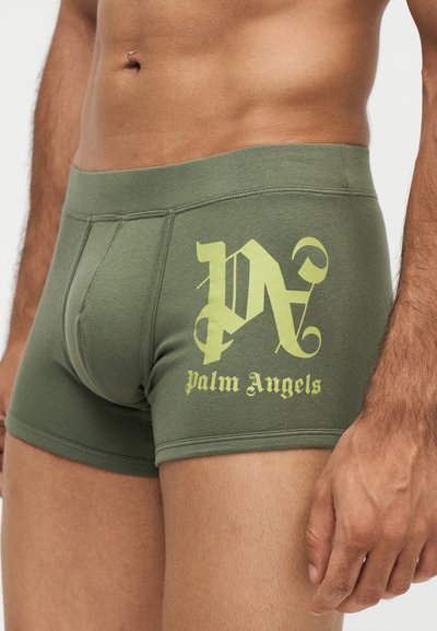 Gros plan d’un homme portant un boxer vert olive avec le logo "Palm Angels" imprimé en jaune sur la cuisse gauche.