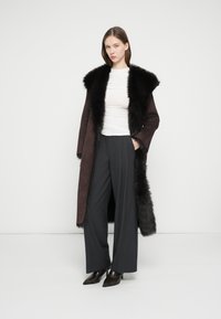 Μακρύ καφέ παλτό από σουέτ με μαύρο γούνινο γιακά, συνδυασμένο με γκρι παντελόνι φαρδύ και μαύρα μυτερά παπούτσια. Λευκό εφαρμοστό τοπ από κάτω.