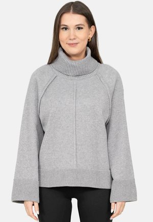 Donna con lunghi capelli castani che indossa orecchini a cerchio d'oro e un maglione dolcevita grigio chiaro con ampi polsini e pantaloni neri.