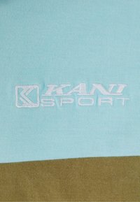 Gaiši zils kokvilnas audums ar gludu tekstūru, ar izšūtu "KANI SPORT" logotipu baltā krāsā un tumši brūnu horizontālu joslu apakšā.
