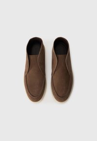 Par de zapatos de ante marrón sin cordones con suelas blancas, vistos desde arriba sobre un fondo blanco liso.