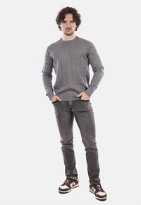 Maglione grigio a maglia con maniche a coste, abbinato a jeans scuri e sneakers marrone e bianco, in piedi davanti a uno sfondo bianco.