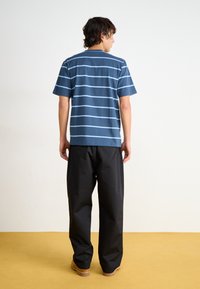 T-shirt rayé bleu marine à manches courtes et col rond, associé à un pantalon large noir, se tenant sur un sol jaune.