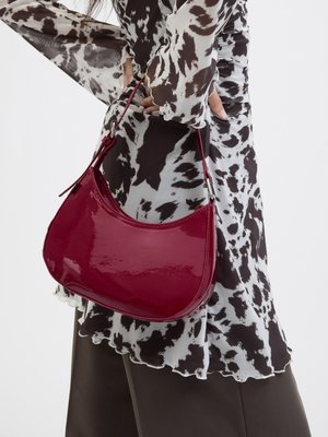 Vrouw met een zwart-wit gebloemde blouse houdt een glanzende rode schoudertas vast tegen een effen achtergrond.