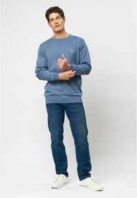 Blå crewneck sweatshirt med ribbede manchetter og kant, parret med mørkeblå jeans og hvide sneakers. Modellen står foran en neutral baggrund.