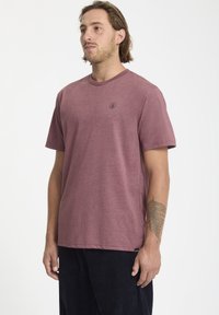 Camiseta de manga corta color burdeos en una tela suave y texturizada, con un cuello redondo y un pequeño detalle del logo en el lado izquierdo del pecho. Combinada con pantalones oscuros.