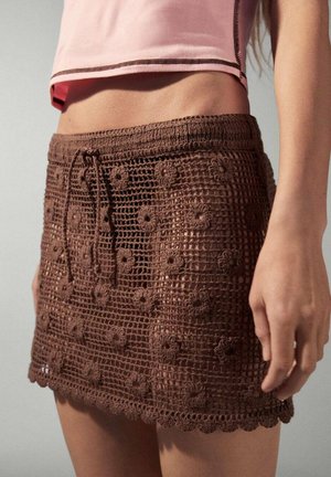 Mini-jupe marron en crochet avec des motifs floraux, portée avec un top court rose laissant apparaitre le ventre et une partie du bras.