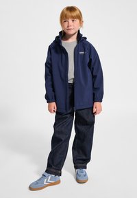 Marinblå vattentät jacka med huva, dragkedjeförslutning, elastiska ärmslut, grå T-shirt, lösa mörka jeans och ljusblå sneakers med vita detaljer.