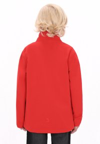 Rød softshell-jakke med lange ærmer, høj krave og en glat tekstur. Har et logo ved kanten og en struktureret pasform.