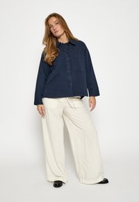 Tumši zils pogājams krekls ar divām krūšu kabatām, kombinēts ar brīviem baltajiem platajām biksēm. Melni apavi pabeidz apģērbu.