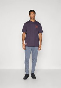 Vijolična bombažna majica s kratkimi rokavi, na prsih rdeč logotip. Kombinirana s svetlo sivimi trenirkami, črnimi supergami in sproščenim krojem.