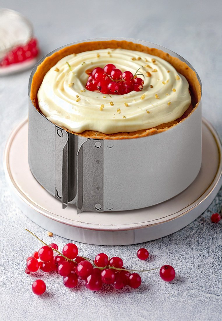 Runder Kuchen mit weißem Sahneguss, garniert mit roten Johannisbeeren, in einem runden metallischen Kuchenteiler. Auf einem hellen Teller platziert, auf dem verstreute rote Johannisbeeren liegen.