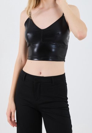 Frau trägt ein schwarzes glänzendes Crop-Top mit dünnen Trägern und schwarze hoch taillierte Hosen, dargestellt vom Hals bis zu den Hüften vor einem einfarbigen Hintergrund.
