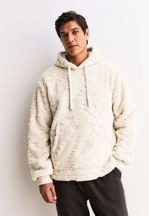 Mężczyzna w beżowej, teksturowanej bluzie z kapturem i kieszenią z przodu oraz w czarnych spodniach, stojący na tle jednolitego białego tła.