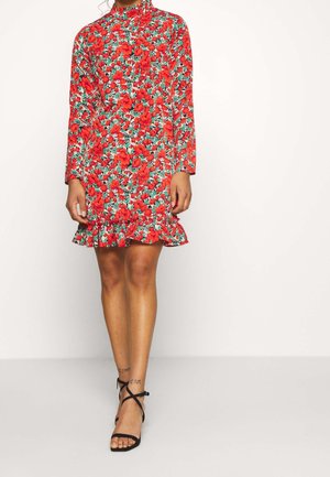 Personne portant une robe fleurie à manches longues et col montant avec des fleurs rouges et des feuilles vertes, associée à des talons noirs avec bride à la cheville.
