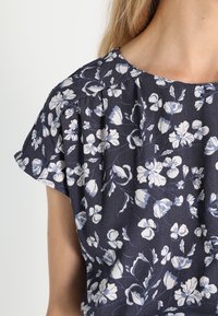 Nahaufnahme einer Person, die eine dunkelblaue Bluse mit weißen und hellblauen Blumenmustern und kurzen, leicht puffigen Ärmeln trägt.