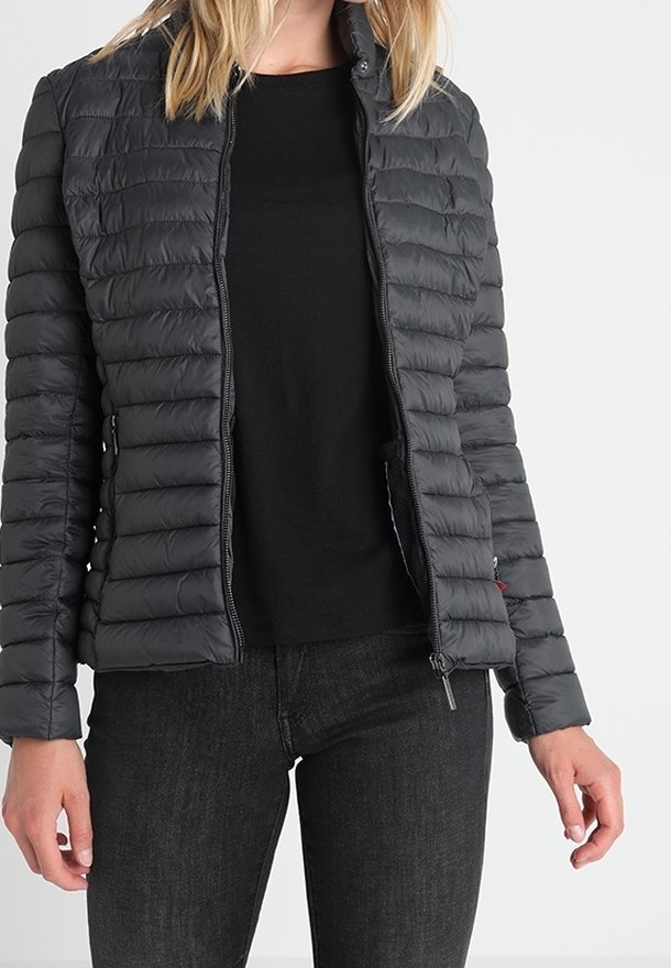 Giacca imbottita nera con motivo trapuntato orizzontale, chiusura con zip e collo alto. Indossata sopra una camicia nera e abbinata a jeans scuri.