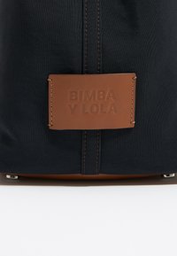 Czarna torba na ramię z tkaniny, z brązową skórzaną metką wytłoczoną napisem "BIMBA Y LOLA", detalami w szyciu oraz metalowymi akcentami u podstawy.