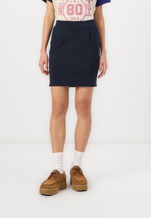 Gonna fitted in cotone blu navy con due tasche frontali, abbinata a una t-shirt grafica beige e scarpe con zeppa marroni. Stylisticamente completata con calzini bianchi.