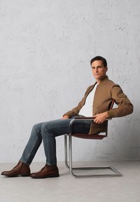 Brązowa kurtka bombowca, biała koszulka, niebieskie dżinsy i brązowe skórzane buty, siedzący na nowoczesnym krześle z chromowanym stelażem na tle o textured.