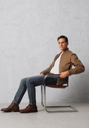 Brązowa kurtka bombowca, biała koszulka, niebieskie dżinsy i brązowe skórzane buty, siedzący na nowoczesnym krześle z chromowanym stelażem na tle o textured.