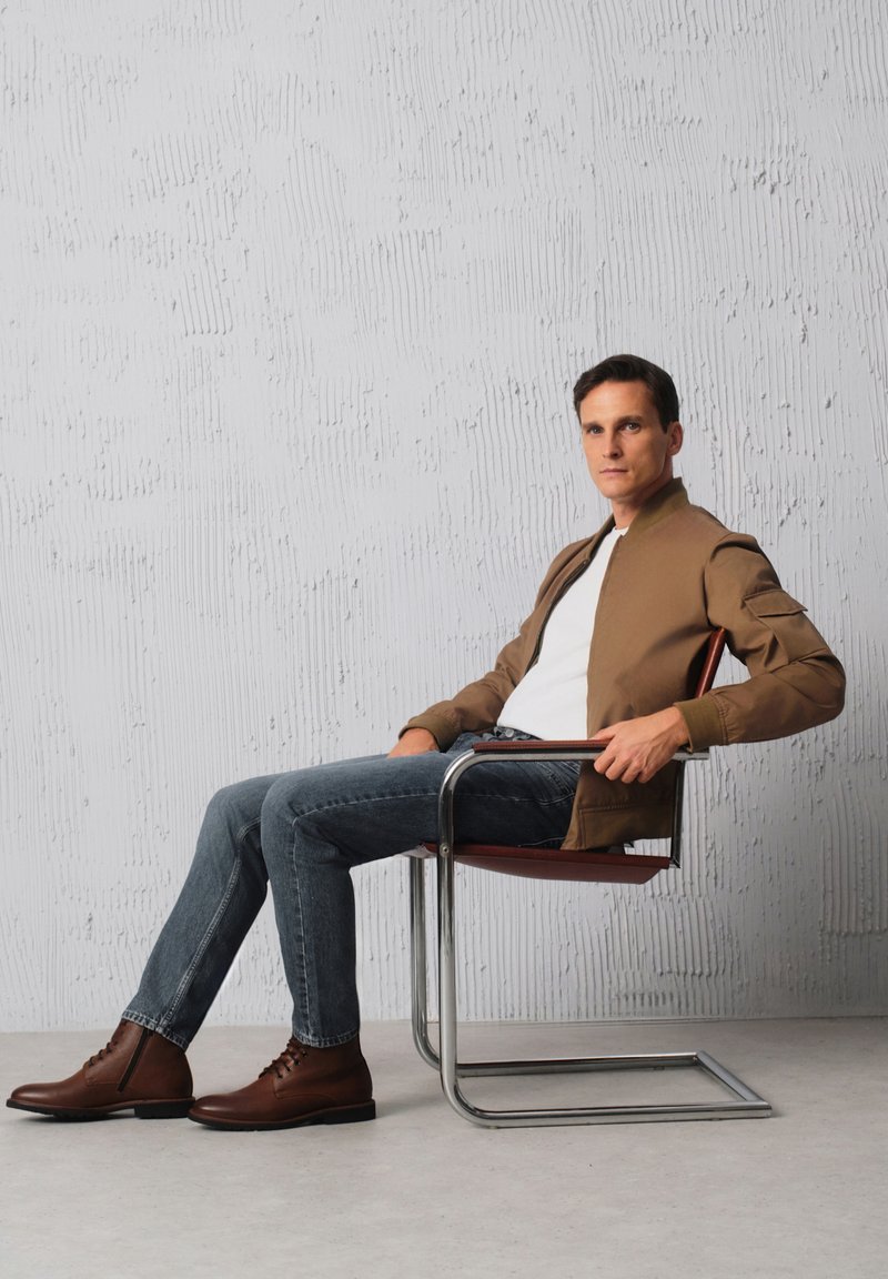 Brązowa kurtka bombowca, biała koszulka, niebieskie dżinsy i brązowe skórzane buty, siedzący na nowoczesnym krześle z chromowanym stelażem na tle o textured.