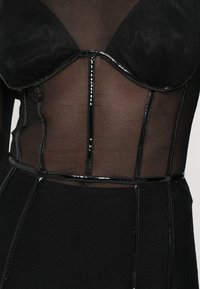 Juodos tinklinės bodice su struktūrinėmis taurėmis, blizgiomis juodomis siūlėmis ir kontrastingomis tekstūromis. Pasižymi prigludusia siluetu ir apibrėžta liemenėlė.